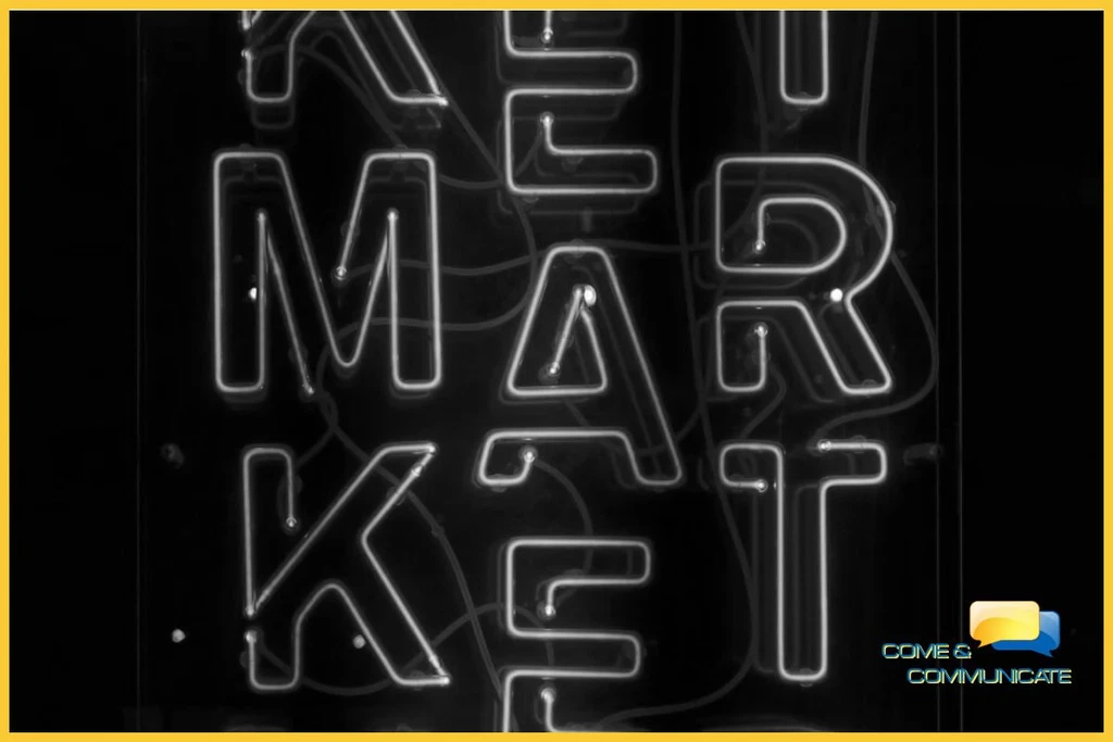 Imagen art&iacute;stica en blanco y negro con un borde amarillo que muestra un letrero de ne&oacute;n brillante con la palabra 'MARKET' dispuesta verticalmente. El dise&ntilde;o tiene un estilo retro-moderno con cables visibles sobre un fondo oscuro, y el logotipo de Come & Communicate aparece en la esquina inferior derecha.