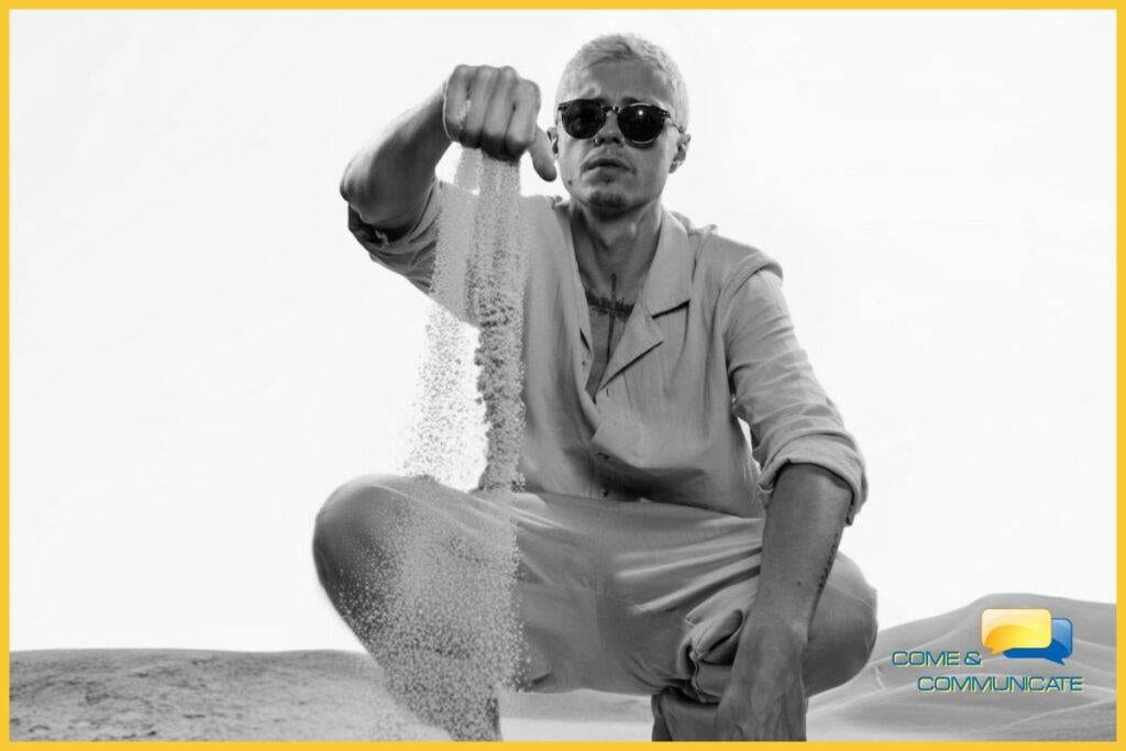 Chico con gafas de sol de cuclillas en un desierto levantando el puño derecho y dejando caer arena de la mano