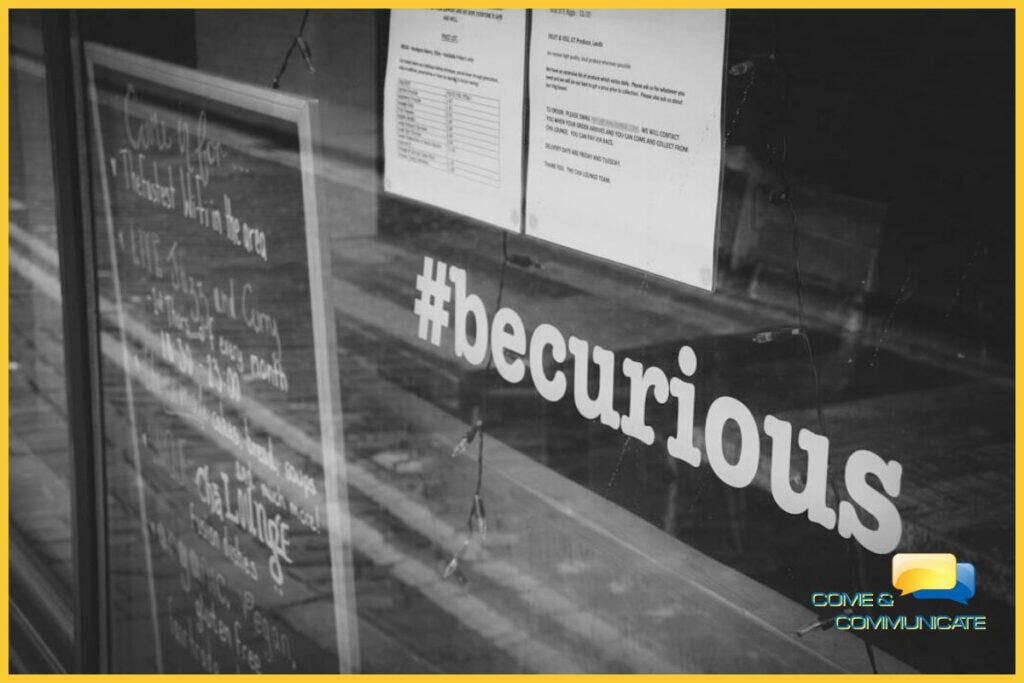 Frase #becurious puesta en vinilo en la ventana de un local junto con una pizarra negra.