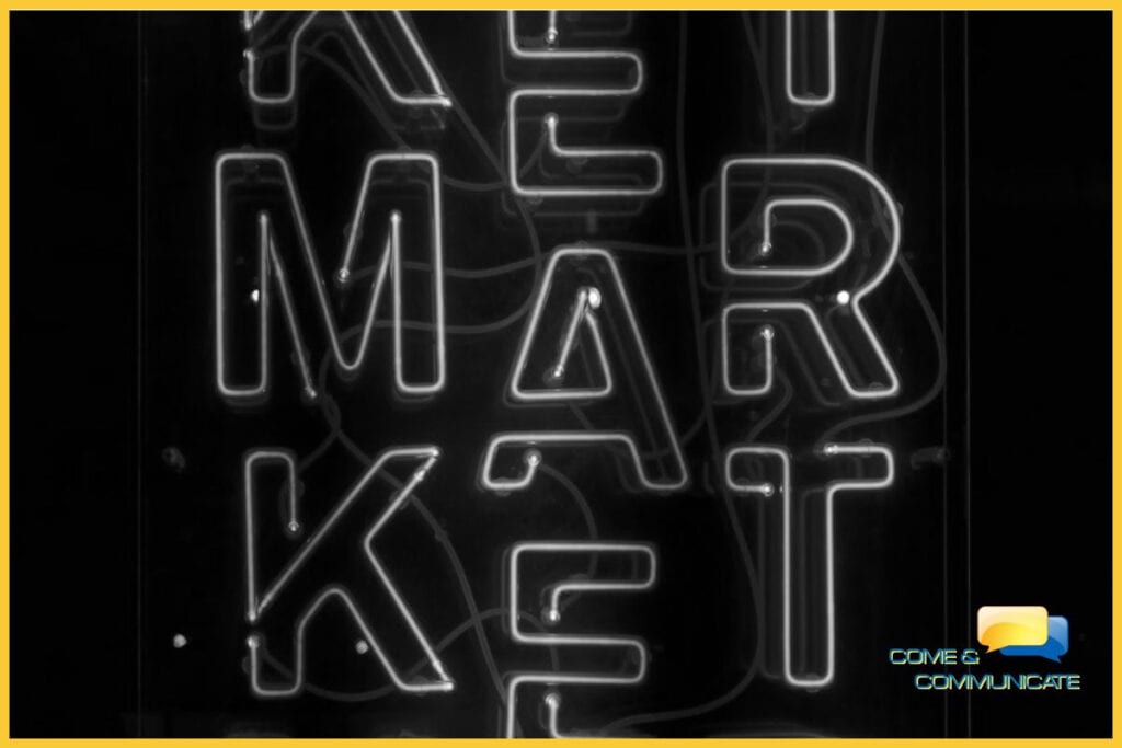 Imagen artística en blanco y negro con un borde amarillo que muestra un letrero de neón brillante con la palabra 'MARKET' dispuesta verticalmente. El diseño tiene un estilo retro-moderno con cables visibles sobre un fondo oscuro, y el logotipo de Come & Communicate aparece en la esquina inferior derecha.
